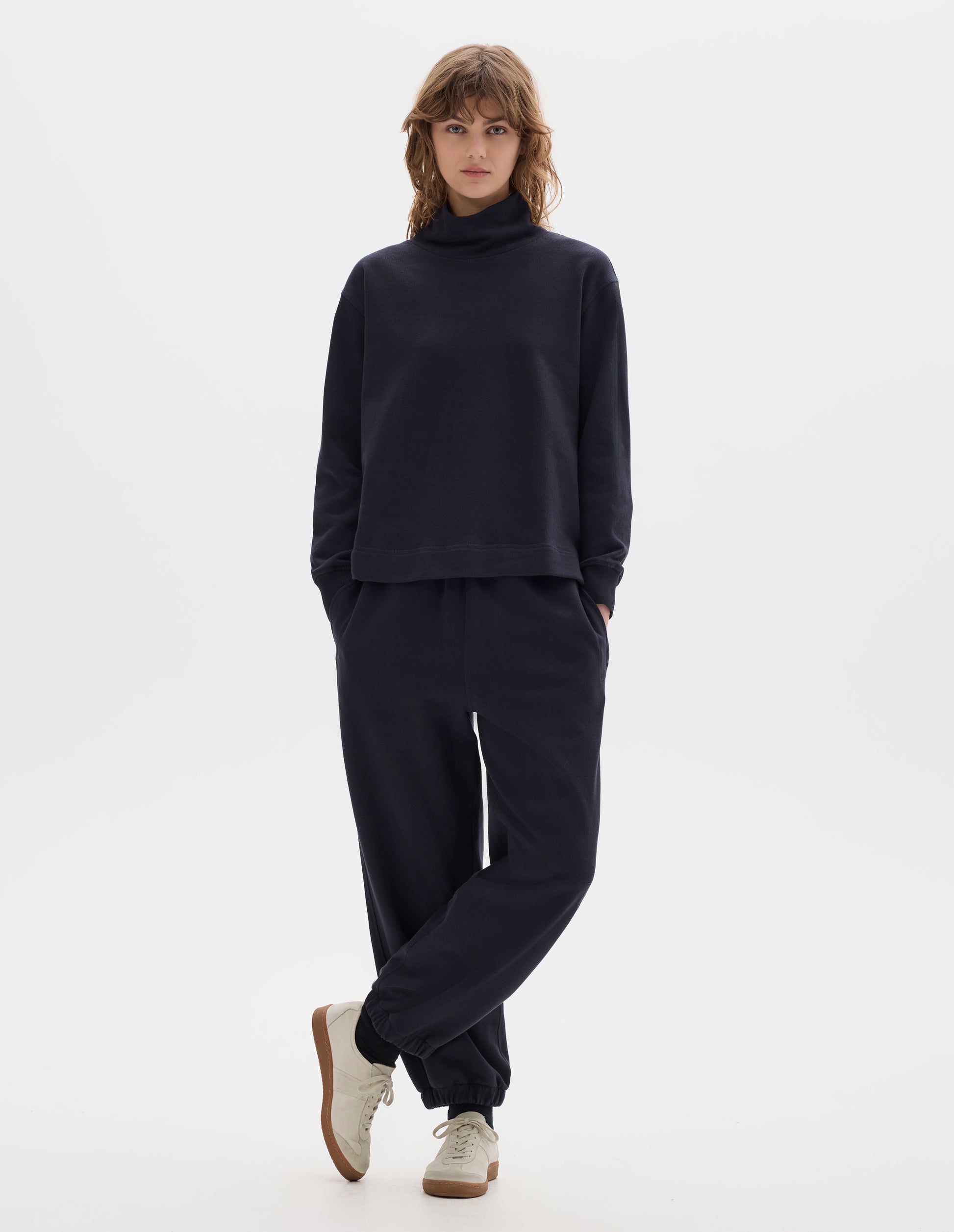 Full body image, front view of model wearing a dark navy loose funnel neck sweatshirt with single rib on cuffs and flatlock stitching on seams. Styled with a dark navy jogger style trousers, black socks and off white suede trainers.