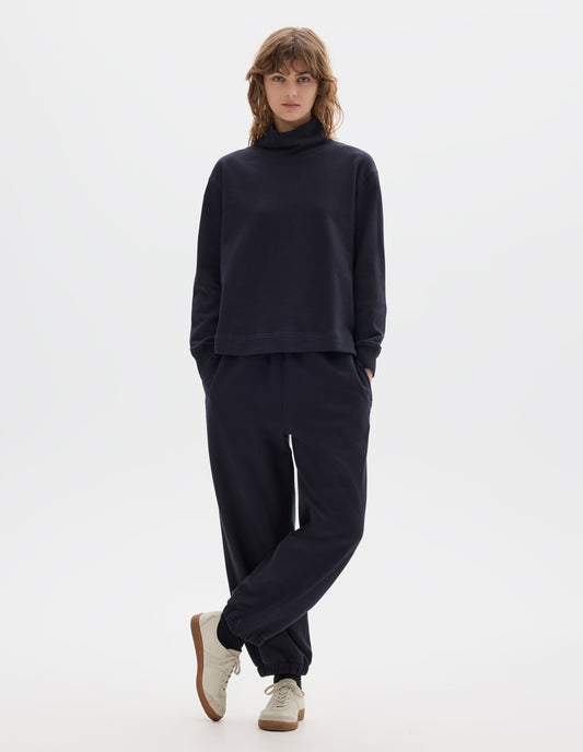 Full body image, front view of model wearing a dark navy loose funnel neck sweatshirt with single rib on cuffs and flatlock stitching on seams. Styled with a dark navy jogger style trousers, black socks and off white suede trainers.