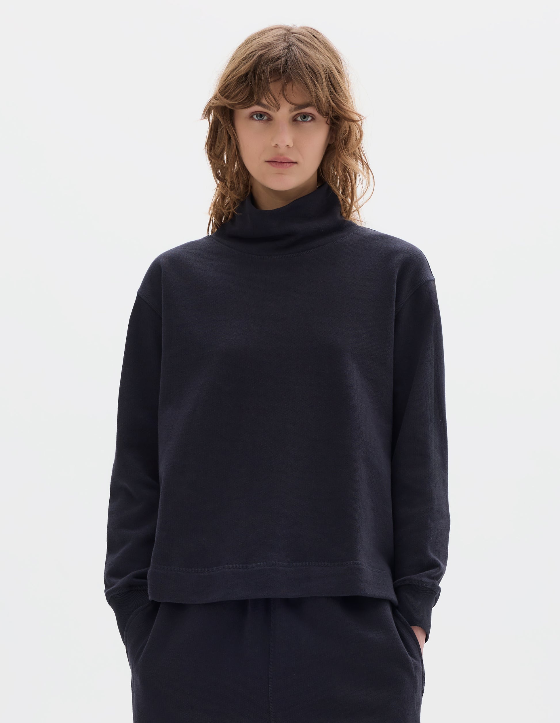 Cropped body image, front view of model wearing a dark navy loose funnel neck sweatshirt with single rib on cuffs and flatlock stitching on seams. Styled with a dark navy jogger style trousers.