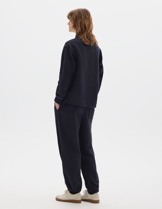 Full body image, back view of model wearing a dark navy loose funnel neck sweatshirt with single rib on cuffs and flatlock stitching on seams. Styled with a dark navy jogger style trousers, black socks and off white suede trainers.