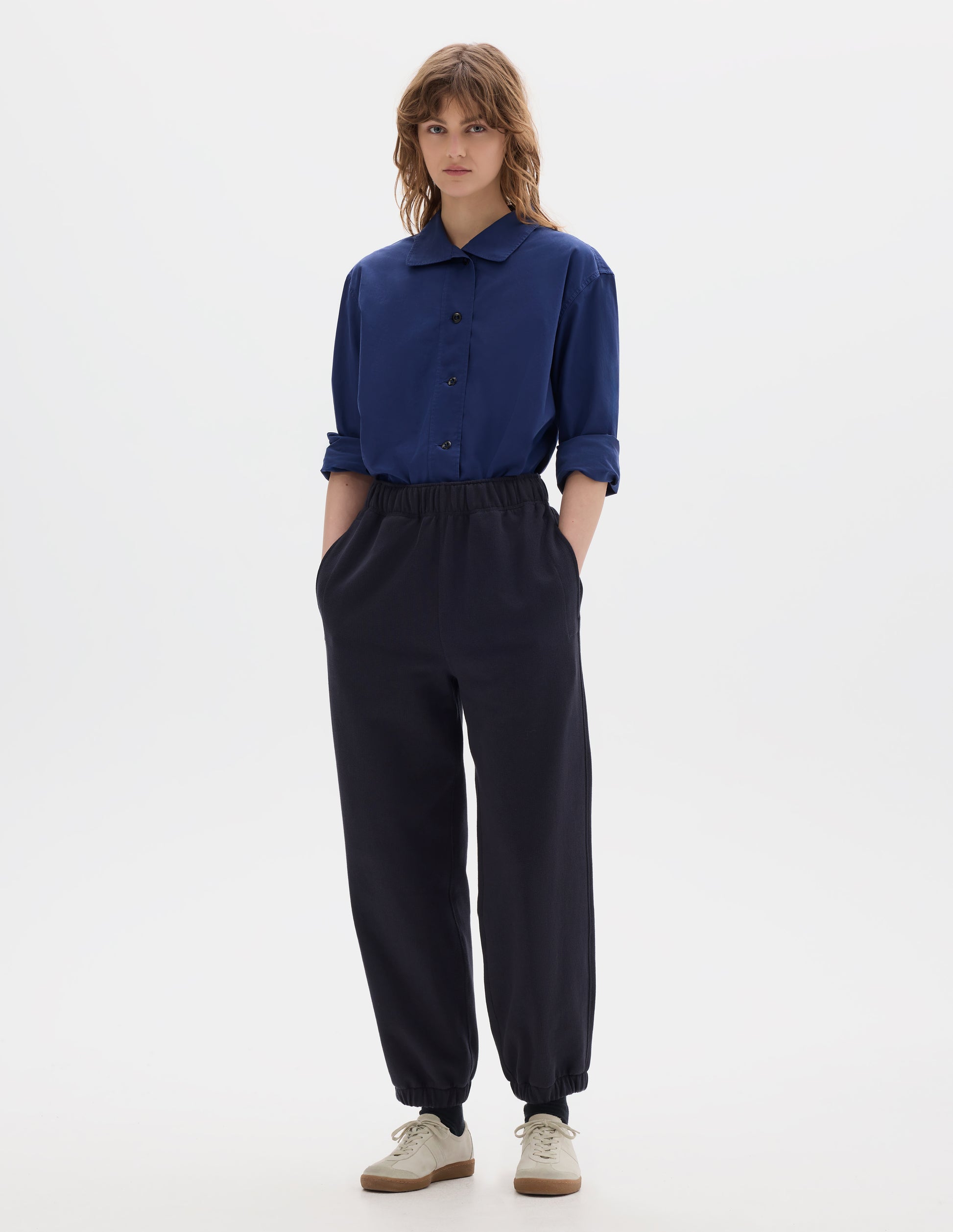 Full body image, front view of model wearing an asymmetric blue cotton shirt, with collar with a softly rounded point and dark buttons. Styled with lightweight jogger style trousers, with side pockets and elasticated waistband and hem. Worn with black socks and off white suede trainers.