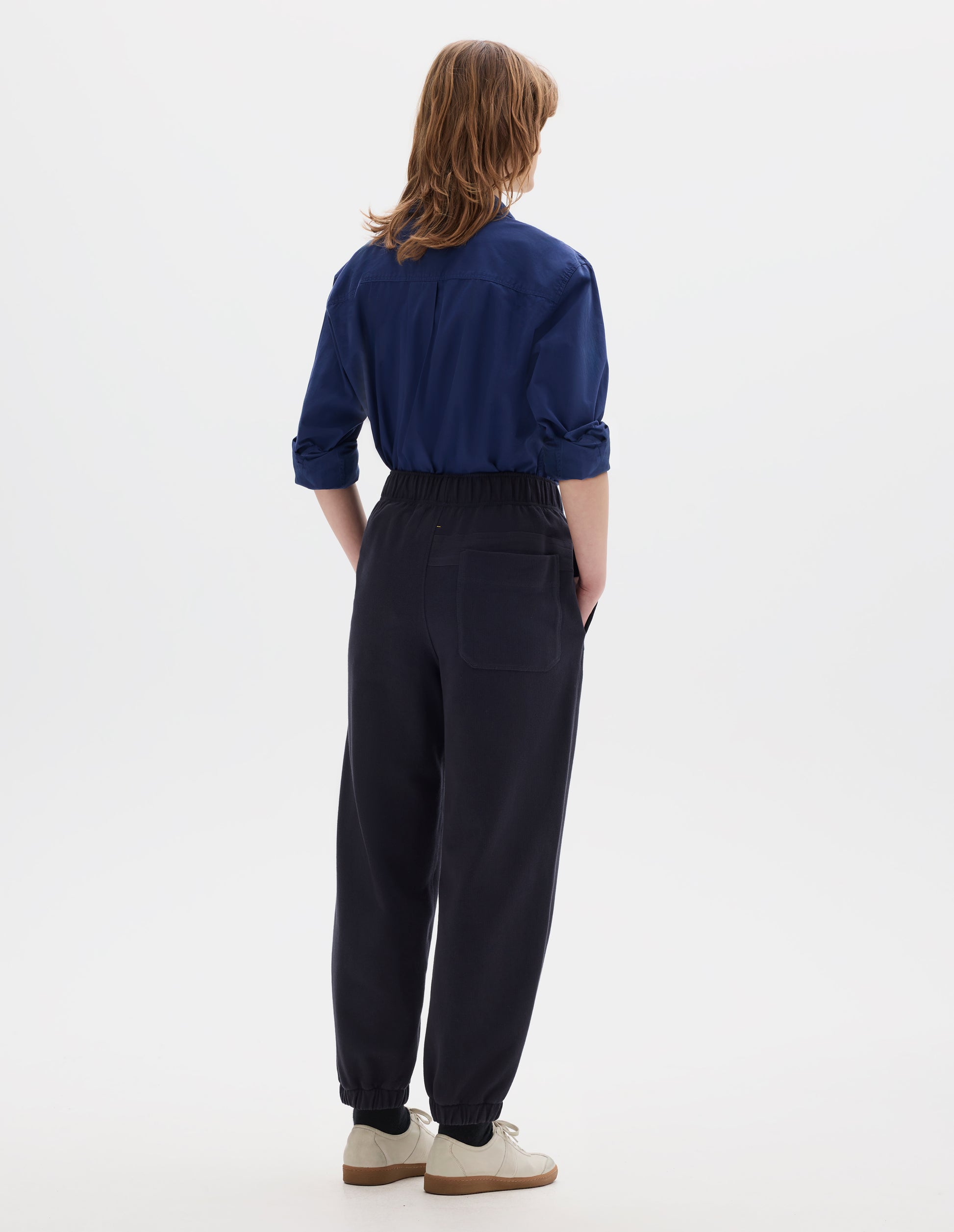 Full body image, back view of model wearing a blue cotton shirt, with box pleat on back yoke. Styled with lightweight jogger style trousers, with back patch pocket and elasticated waistband and hem. Worn with black socks and off white suede trainers.