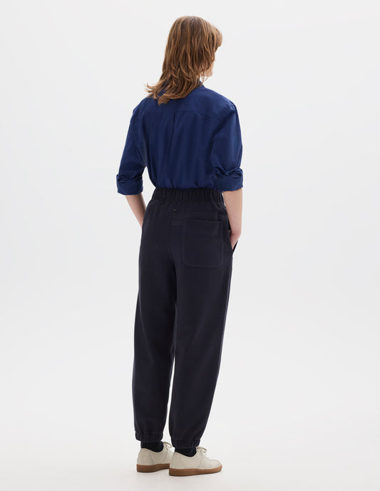 Full body image, back view of model wearing a blue cotton shirt, with box pleat on back yoke. Styled with lightweight jogger style trousers, with back patch pocket and elasticated waistband and hem. Worn with black socks and off white suede trainers.