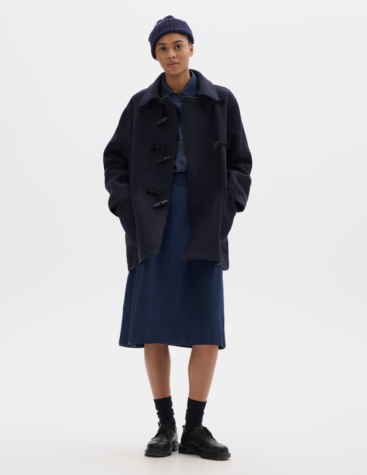 Full body image, front view of model wearing a mid length dark blue coat with wooden toggles fastening and patch pockets, worn over a blue shirt and styled with a dark blue ribbed beanie, fingerless gloves, blue knee length pleated skirt, black socks and leather shoes.