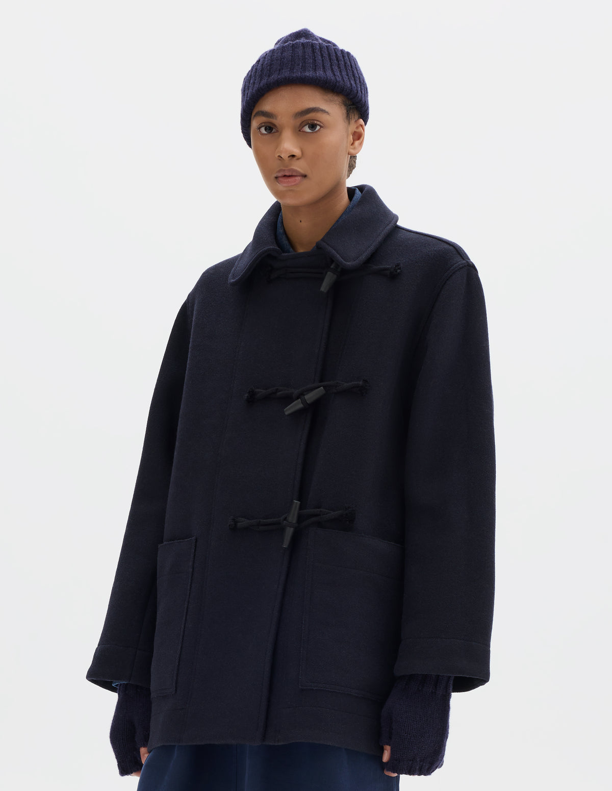 Cropped body image, front view of model wearing a mid length dark blue coat with wooden toggles fastening and patch pockets, worn over a blue shirt and styled with a dark blue ribbed beanie, fingerless gloves and a blue pleated skirt.
