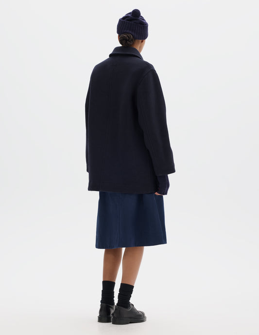 Full body image, back view of model wearing a mid length dark blue coat with a dropped shoulder, styled with a dark blue ribbed beanie with a pom pom on top, fingerless gloves, blue knee length skirt, black socks and leather shoes.
