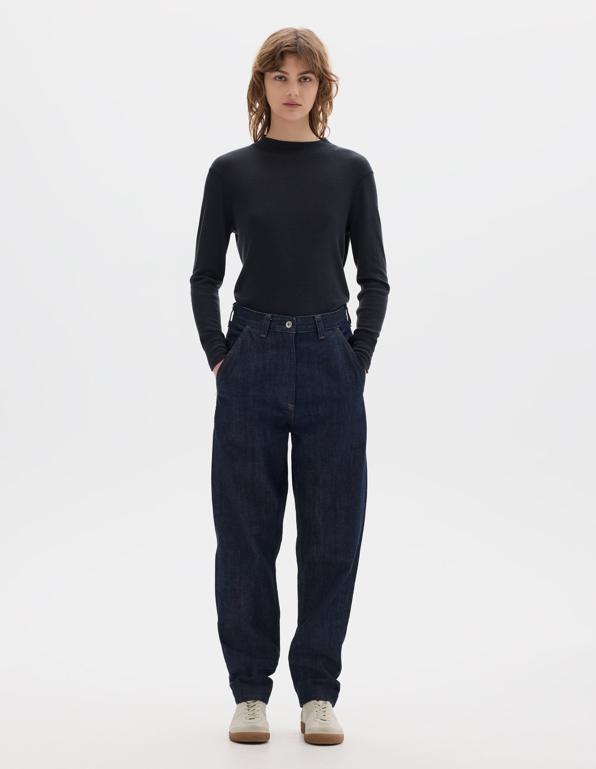 Full body image, front view of model wearing  relaxed dark blue jeans with slant side pockets. Styled with an off black long sleeve thermal t shirt and beige suede trainers.