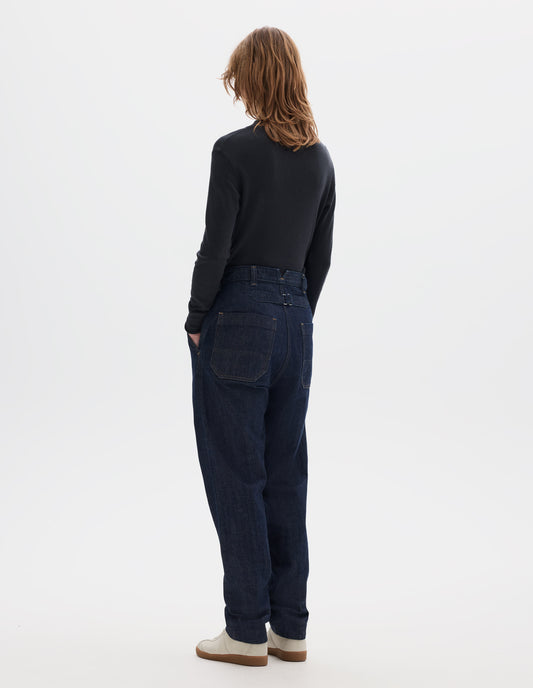 Full body image, back view of model wearing relaxed dark blue jeans with large patch back pockets and cinch on back. Styled with an off black long sleeve thermal t shirt and beige suede trainers.