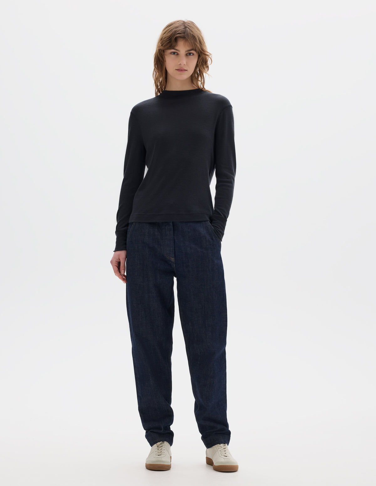 Full body image, front view of model wearing black long sleeve t shirt with set-in sleeves, a rib neck and cuffs with flatlock stitching. Styled with indigo denim jeans and white trainers with gum sole. 