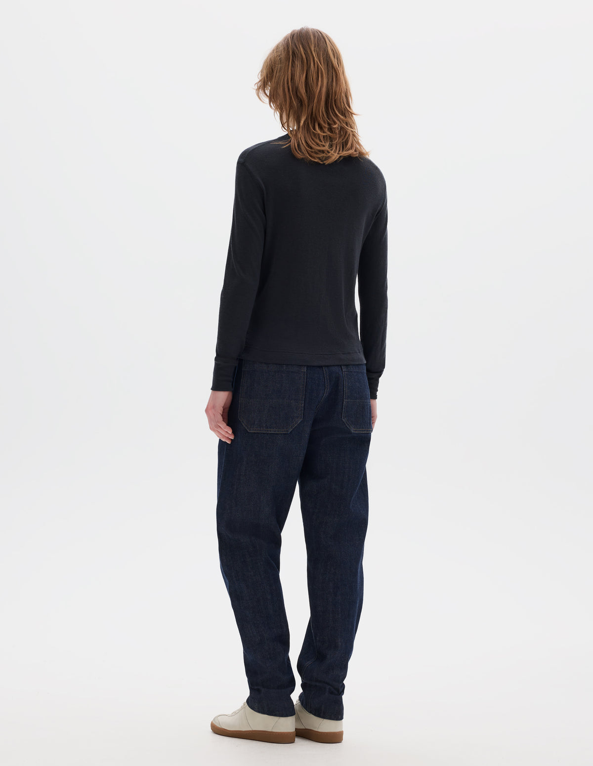 Full body image, back view of model wearing black long sleeve t shirt with set-in sleeves, a rib neck and cuffs with flatlock stitching. Styled with indigo denim jeans and white trainers with gum sole. 