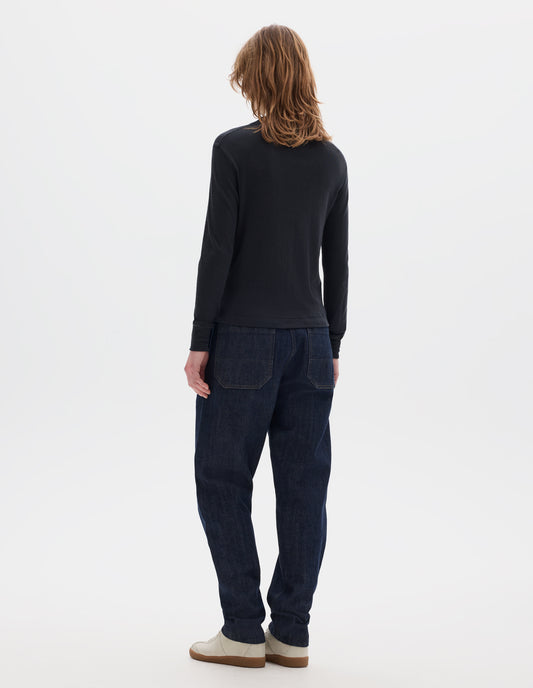 Full body image, back view of model wearing black long sleeve t shirt with set-in sleeves, a rib neck and cuffs with flatlock stitching. Styled with indigo denim jeans and white trainers with gum sole. 
