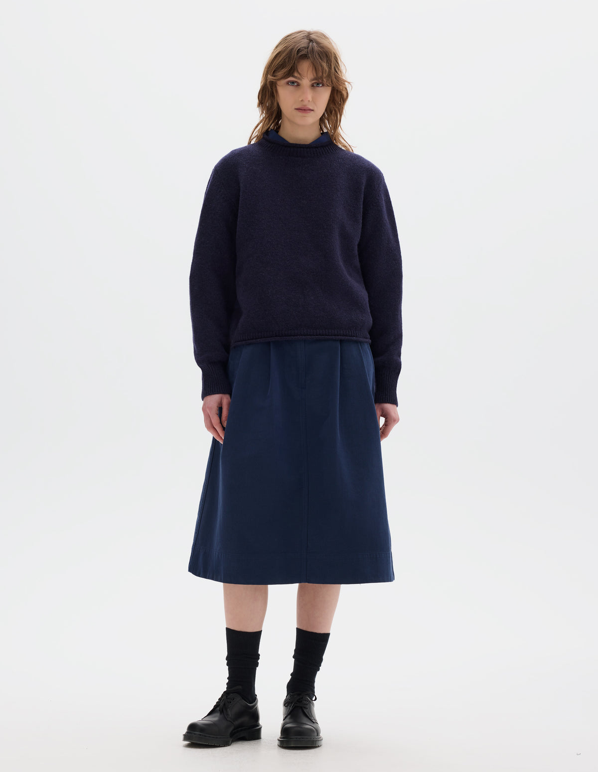 Full body image, front view of model wearing a dark blue midweight crew neck sweater with rib detailing and a rolled edge at the neck and hem. Worn over a blue shirt and styled with a blue pleated skirt, black socks and leather shoes.