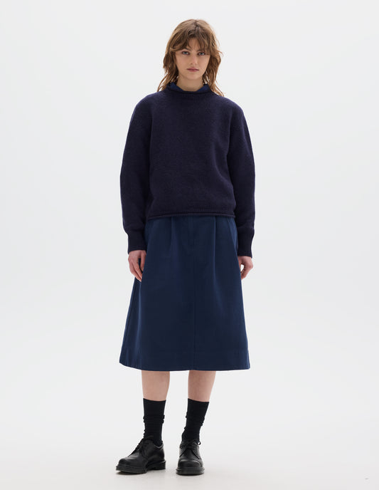 Full body image, front view of model wearing a dark blue midweight crew neck sweater with rib detailing and a rolled edge at the neck and hem. Worn over a blue shirt and styled with a blue pleated skirt, black socks and leather shoes.