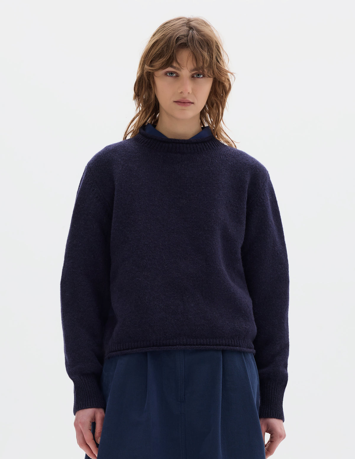 Cropped body image, front view of model wearing a dark blue midweight crew neck sweater with rib detailing and a rolled edge at the neck and hem. Worn over a blue shirt and styled with a blue pleated skirt.