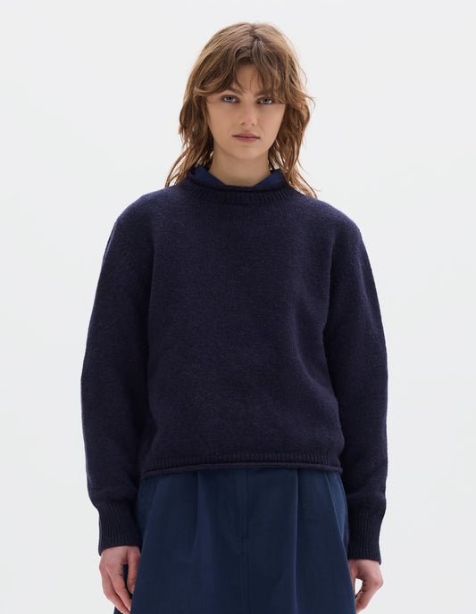 Cropped body image, front view of model wearing a dark blue midweight crew neck sweater with rib detailing and a rolled edge at the neck and hem. Worn over a blue shirt and styled with a blue pleated skirt.