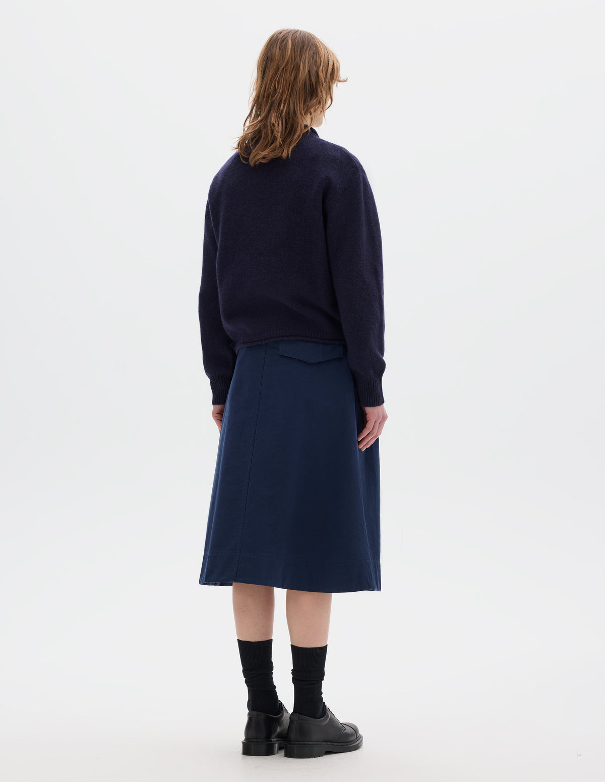 Full body image, back view of model wearing a dark blue midweight crew neck sweater with rib detailing and a rolled edge at the neck and hem. Rib detail on cuffs. Worn over a blue shirt and styled with a blue skirt, black socks and leather shoes.