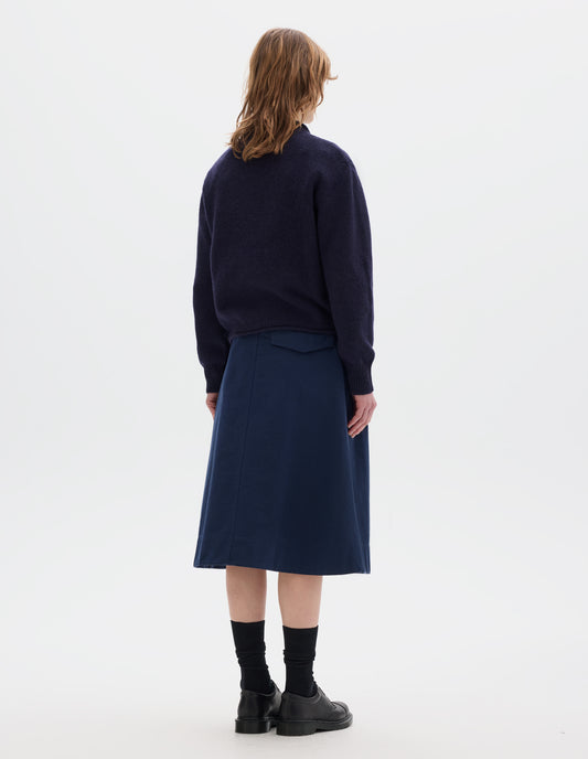 Full body image, back view of model wearing a dark blue midweight crew neck sweater with rib detailing and a rolled edge at the neck and hem. Rib detail on cuffs. Worn over a blue shirt and styled with a blue skirt, black socks and leather shoes.