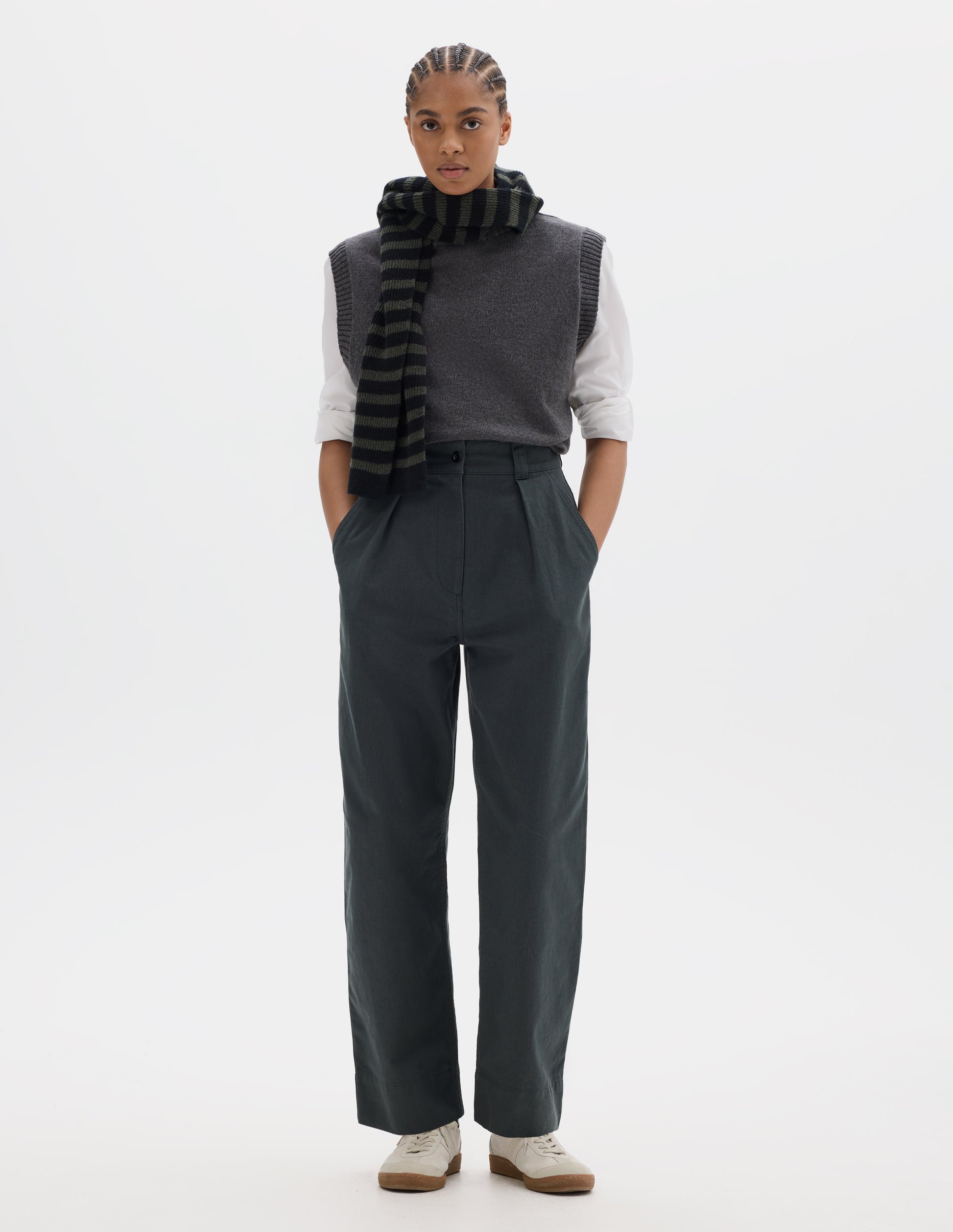 Full body image, front view of model wearing a greyish green high waisted pleated trousers with a single pleat on front. Styled with a grey wool slipover, worn over a white shirt, grey black stripe wool scarf and off white suede trainers.