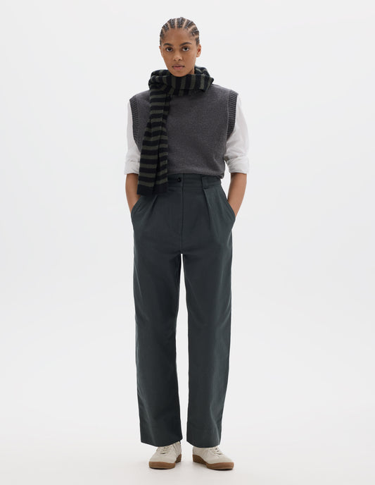 Full body image, front view of model wearing a greyish green high waisted pleated trousers with a single pleat on front. Styled with a grey wool slipover, worn over a white shirt, grey black stripe wool scarf and off white suede trainers.