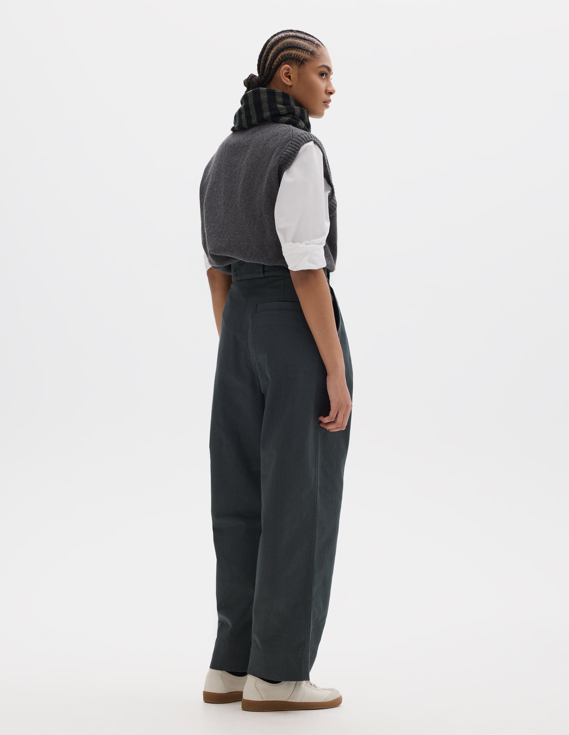 Full body image, back view of model wearing a greyish green high waisted pleated trousers with a stitch-through back welt pocket. Styled with a grey wool slipover, worn over a white shirt, grey black stripe wool scarf and off white suede trainers.
