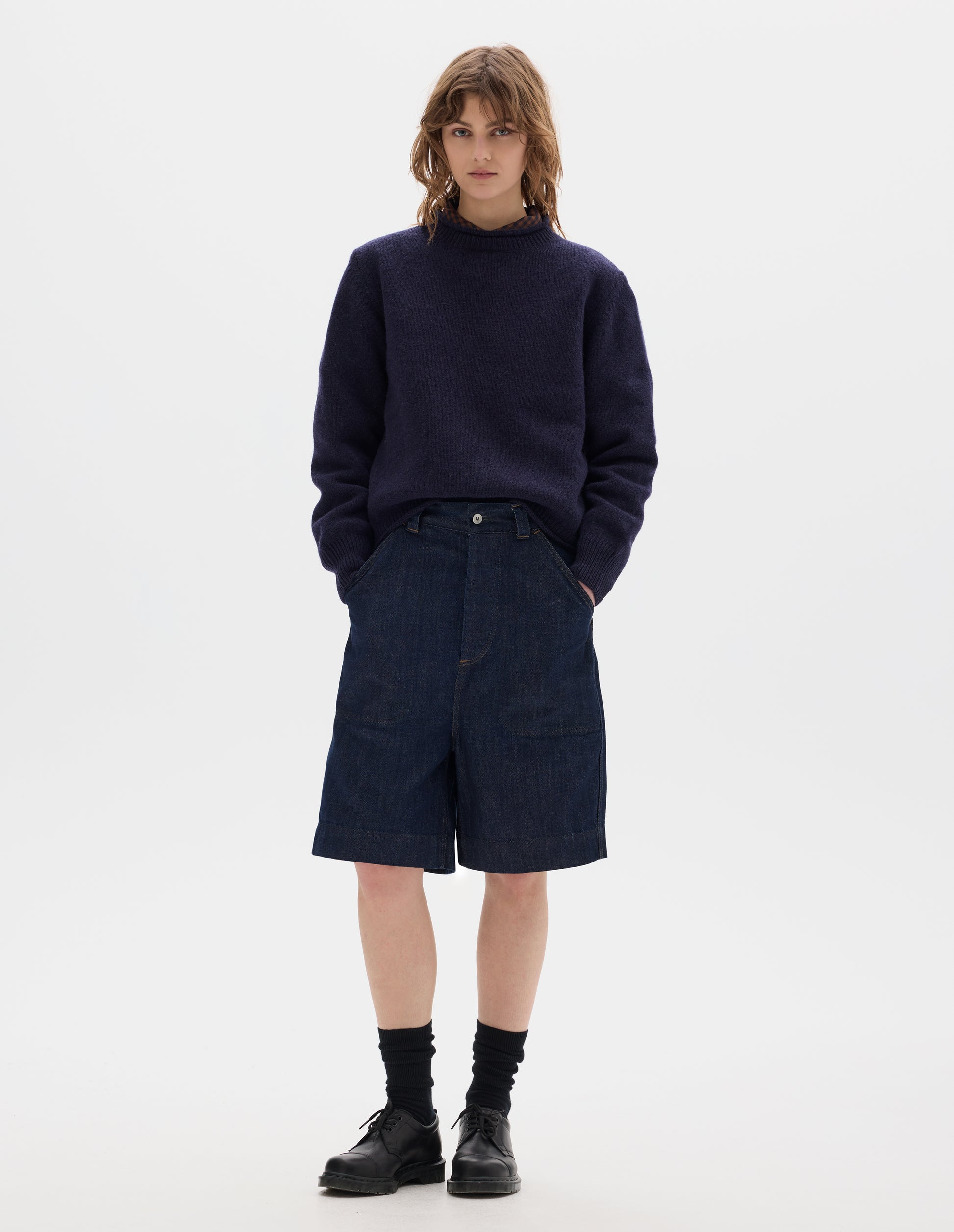 Full body image, front view of model wearing dark blue relaxed denim twill shorts with metal button tab waist adjuster, metal button fly, belt loops and slant stitch-through pockets. Styled with wool navy crew neck jumper, worn over a brown and dark navy gingham shirt, black socks and leather shoes.