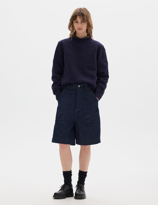 Full body image, front view of model wearing dark blue relaxed denim twill shorts with metal button tab waist adjuster, metal button fly, belt loops and slant stitch-through pockets. Styled with wool navy crew neck jumper, worn over a brown and dark navy gingham shirt, black socks and leather shoes.