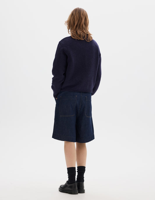 Full body image, back view of model wearing dark blue relaxed denim twill shorts with metal button tab waist adjusters, belt loops and patch pockets. Styled with wool navy crew neck jumper, black socks and leather shoes.