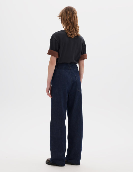Full body image, back view of model wearing indigo blue wide leg light denim twill jeans. With slant side pockets and patch back pocket. Styled with black short sleeve t shirt with faced cuffs with a contrast brown on the reverse and brown leather shoes.