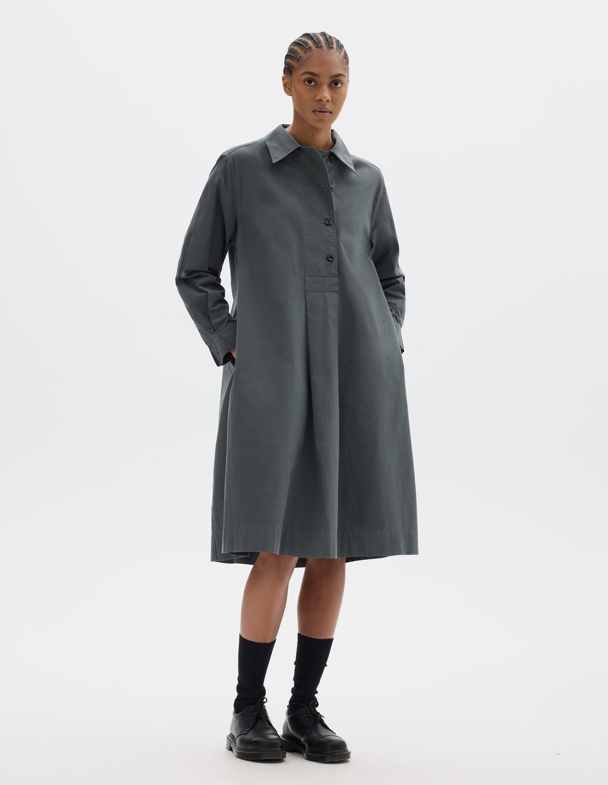 Full body image, front view of model wearing a mid length dark grey pull on cotton dress with a wide offset button placket, a box pleat skirt and side seam pockets. Styled with black socks and leather shoes.