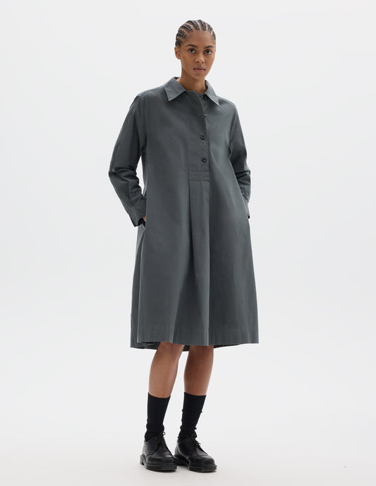 Full body image, front view of model wearing a mid length dark grey pull on cotton dress with a wide offset button placket, a box pleat skirt and side seam pockets. Styled with black socks and leather shoes.