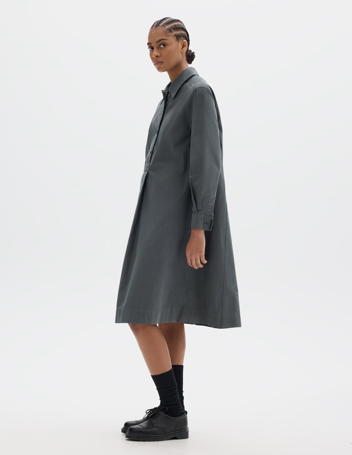 Full body image, front side view of model wearing a mid length dark grey pull on cotton dress with a box pleat skirt and double pleat shirt cuffs. Styled with black socks and leather shoes.