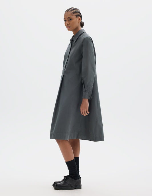 Full body image, front side view of model wearing a mid length dark grey pull on cotton dress with a box pleat skirt and double pleat shirt cuffs. Styled with black socks and leather shoes.