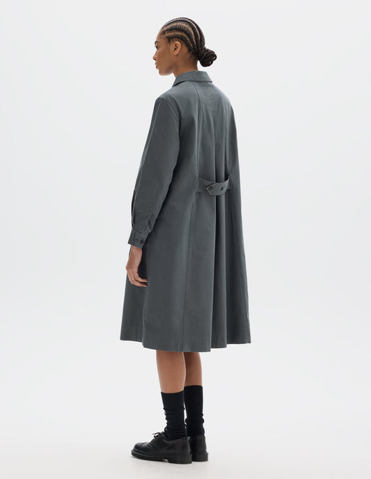 Full body image, back view of model wearing a mid length dark grey pull on cotton dress with an adjustable button belt at the back panel and double pleat shirt cuffs. Styled with black socks and leather shoes.