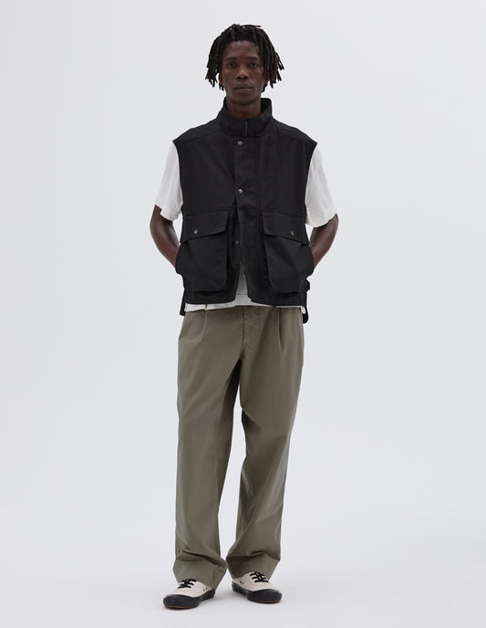 Full body image, front view of model wearing sleeveless black jacket with press stud fastening closure and flap bellow pockets. Styled with white t-shirt, light green trousers and white canvas trainers. 