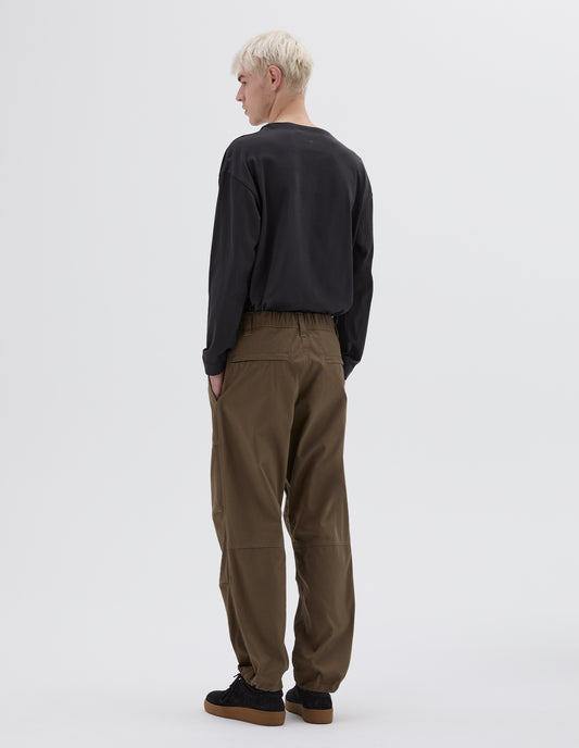 Full body image, back view of model wearing green trousers with elasticated waistband, zip welt back pockets and adjustable drawcord hem. Styled with black long sleeve smock neck t-shirt and black nubuck trainers.