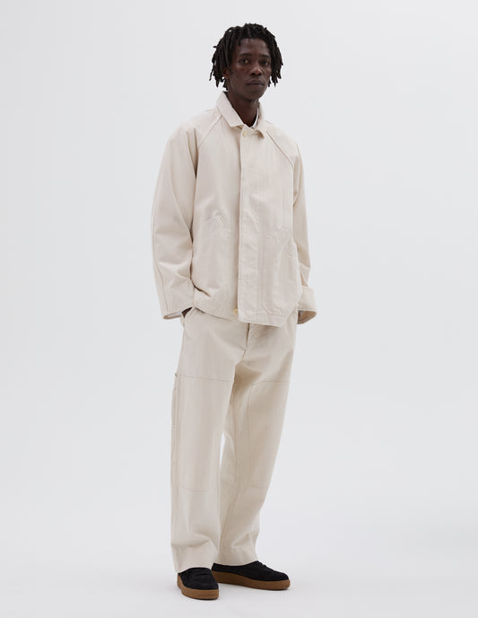 Full body image, front view of model wearing off white raglan jacket with slant patch pockets and corozo buttons. Styled with matching trousers and black nubuck trainers. 