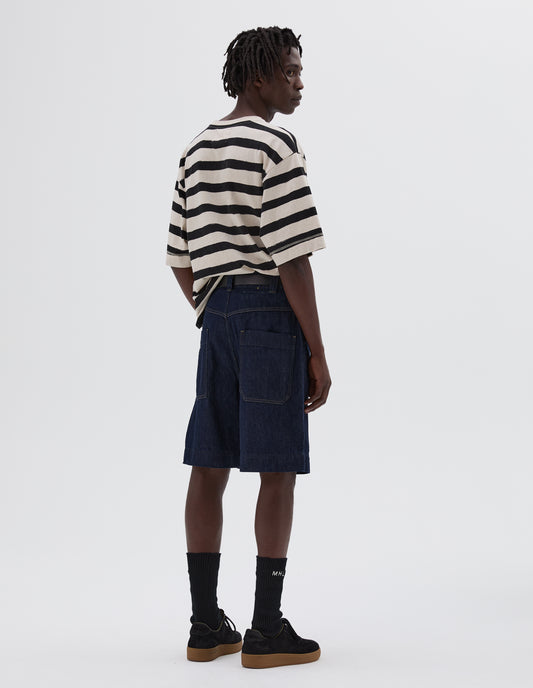 Full body image, back view of model wearing indigo blue denim twill shorts. With metal button tab waist adjusters, belt loops and patch and hammer pocket on back of trousers. Styled with white and black striped t-shirt, black leather belt, black socks and black suede trainers.