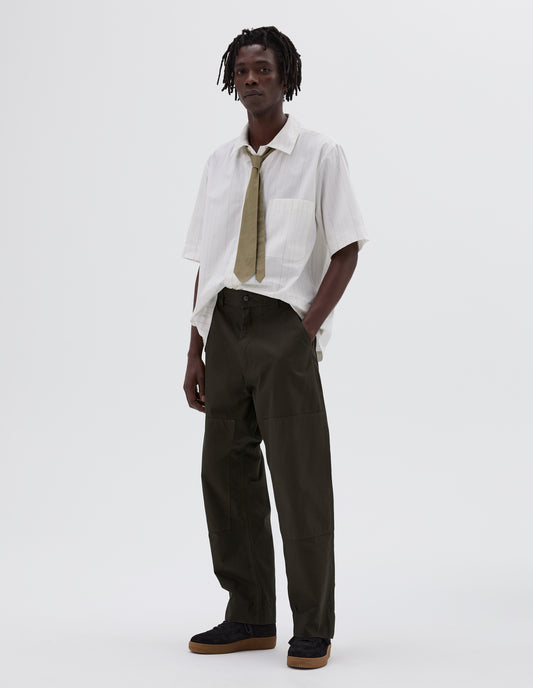 Full body image, side view of model wearing green trousers with large knee patches, double tool pocket on the side leg and zip fly. Styled with short sleeve white shirt, pale green tie and black nubuck trainers.