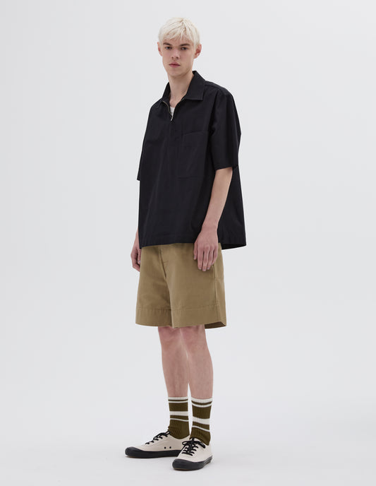 Full body image, front view of model wearing black textured cotton short sleeve shirt, with large patch chest pocket, herringbone detailing on the zip placket, side split hem and elongated back. Styled over white vest, with beige shorts, striped brown and white socks and white canvas trainers.