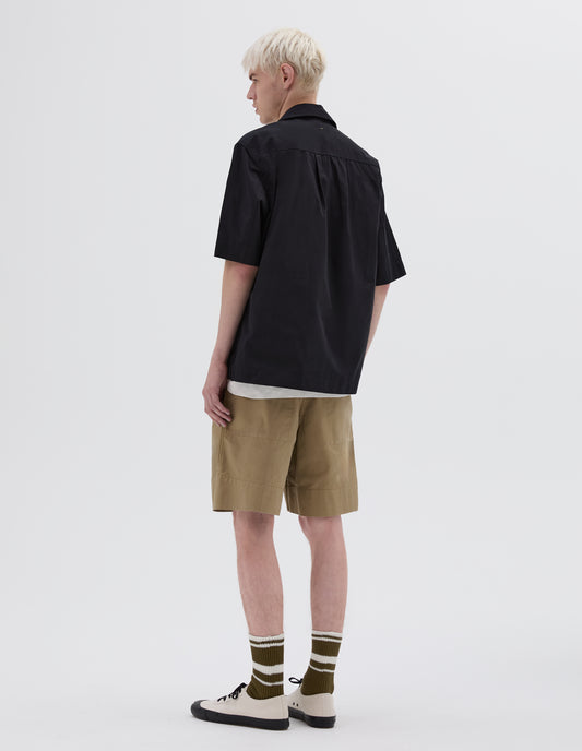 Full body image, back view of model wearing  black textured cotton short sleeve shirt, with side split hem, elongated back and box pleat on back yoke. Styled with beige shorts, striped brown and white socks and white canvas trainers.