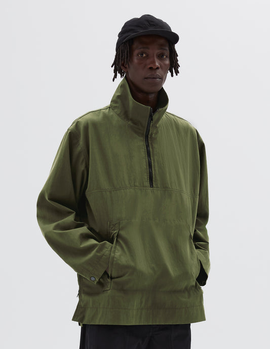 Cropped image, front view of model wearing green anorak with half zip closure, stitch-through flap kangaroo pockets and popper fastening adjustable cuffs. Styled with black trousers and black cap.
