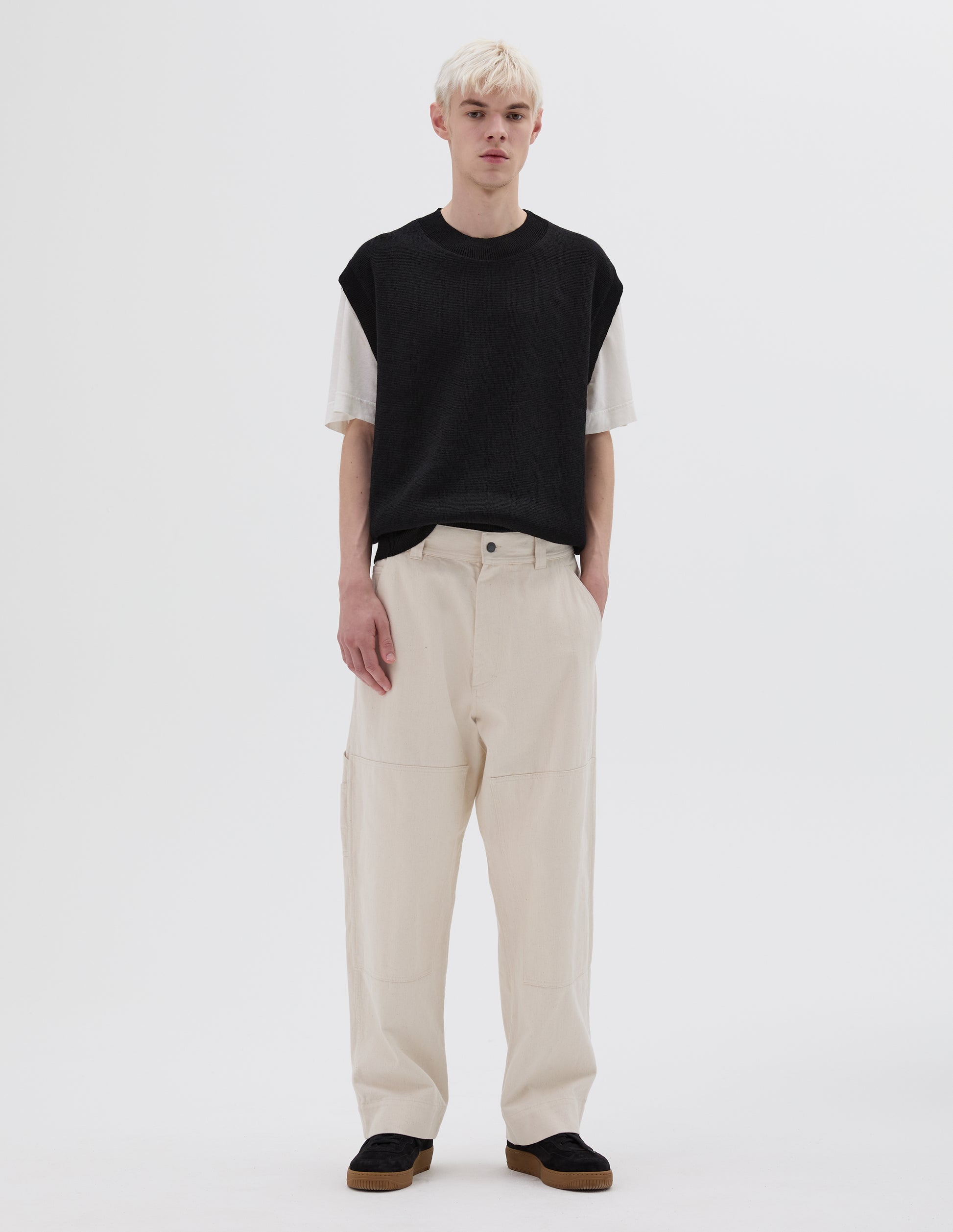 Full body image, front view of model wearing off white trousers with large knee patches, double tool pocket on the side leg and zip fly. Styled with white t-shirt, brown and black sleeveless jumper and black nubuck trainers.