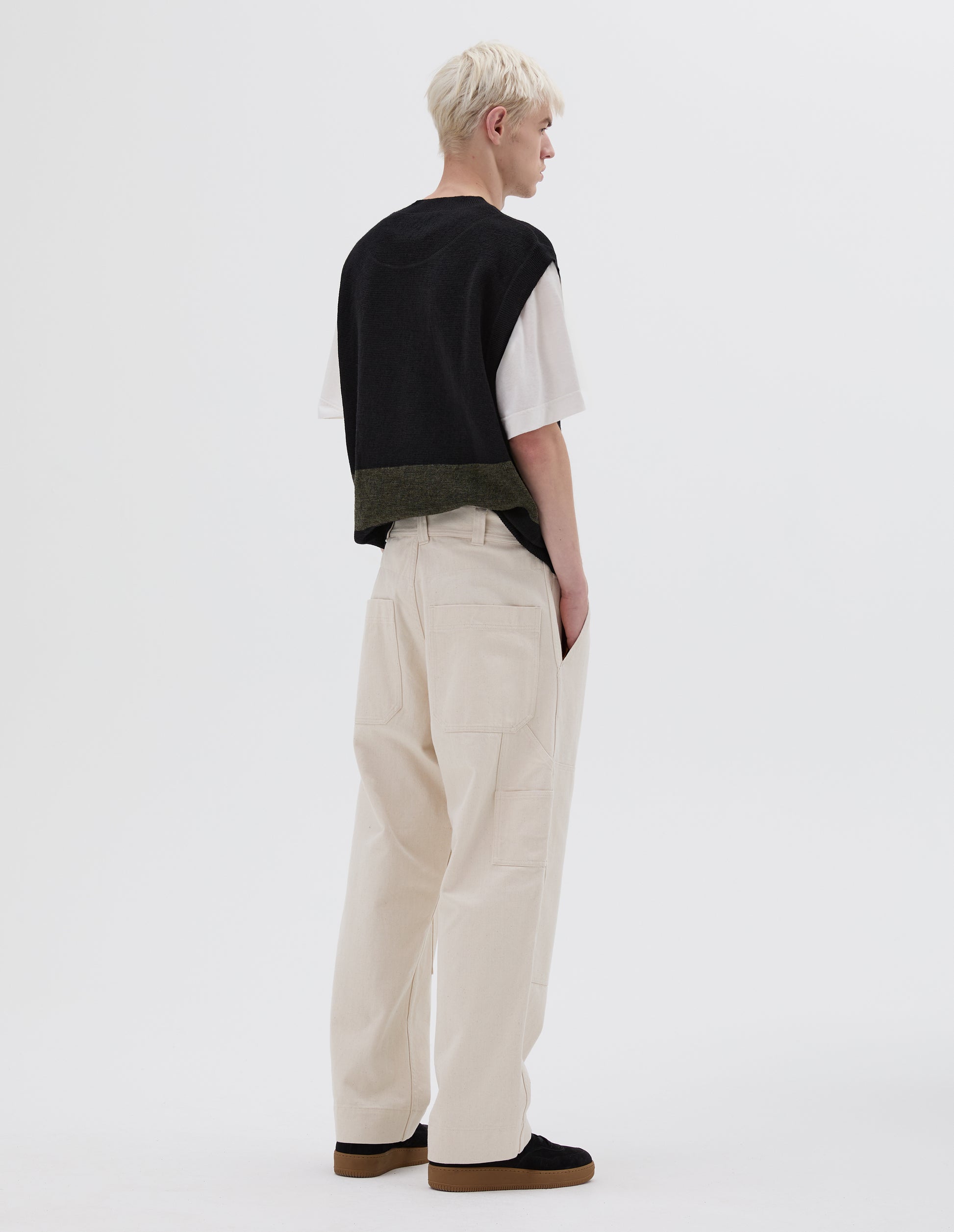 Full body image, back view of model wearing off white trousers with double tool pocket on the side leg, belt loops and large patch back pockets. Styled with white t-shirt, brown and black sleeveless jumper and black nubuck trainers.