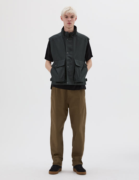 Full body image, front view of model wearing sleeveless dark green jacket with press stud fastening closure and flap bellow pockets. Styled with black t-shirt, green trousers and black nubuck leather trainers.