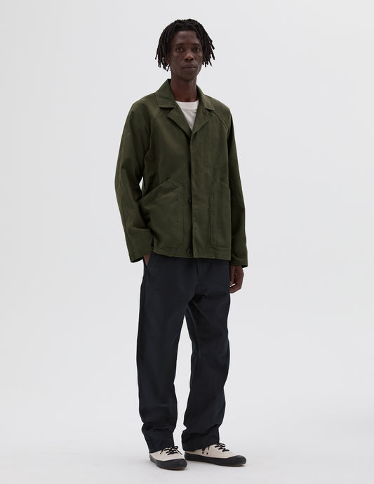 Full body image, front view of model wearing green raglan jacket with slant patch pockets and corozo buttons. Layered over a white t-shirt and worn with navy trousers and white canvas trainers. 