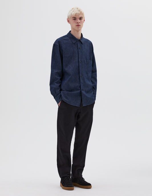 Full body image, front view of model wearing indigo blue long sleeve shirt with raised placket, chest pocket and side gusset stitch detailing. Styled with black trousers and black suede trainers.