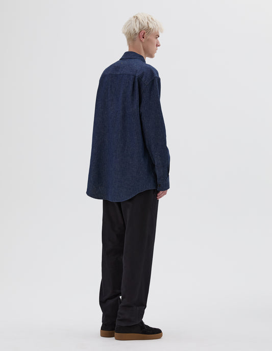 Full body image, back view of model wearing indigo blue long sleeve shirt with side gusset stitch detailing, corozo button cuffs and box pleats on back yoke. Styled with black trousers and black suede trainers.