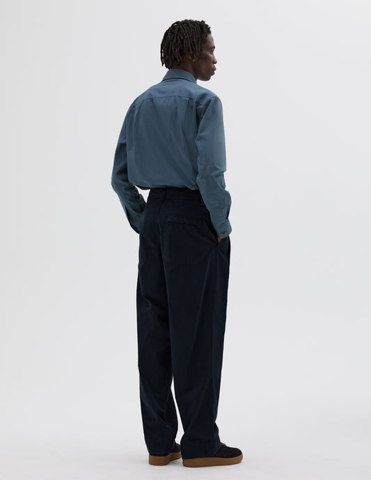 Full body image, back view of model wearing dark navy double pleated trousers with zip fly back pocket. Styled with blue long sleeve shirt and black nubuck trainers.