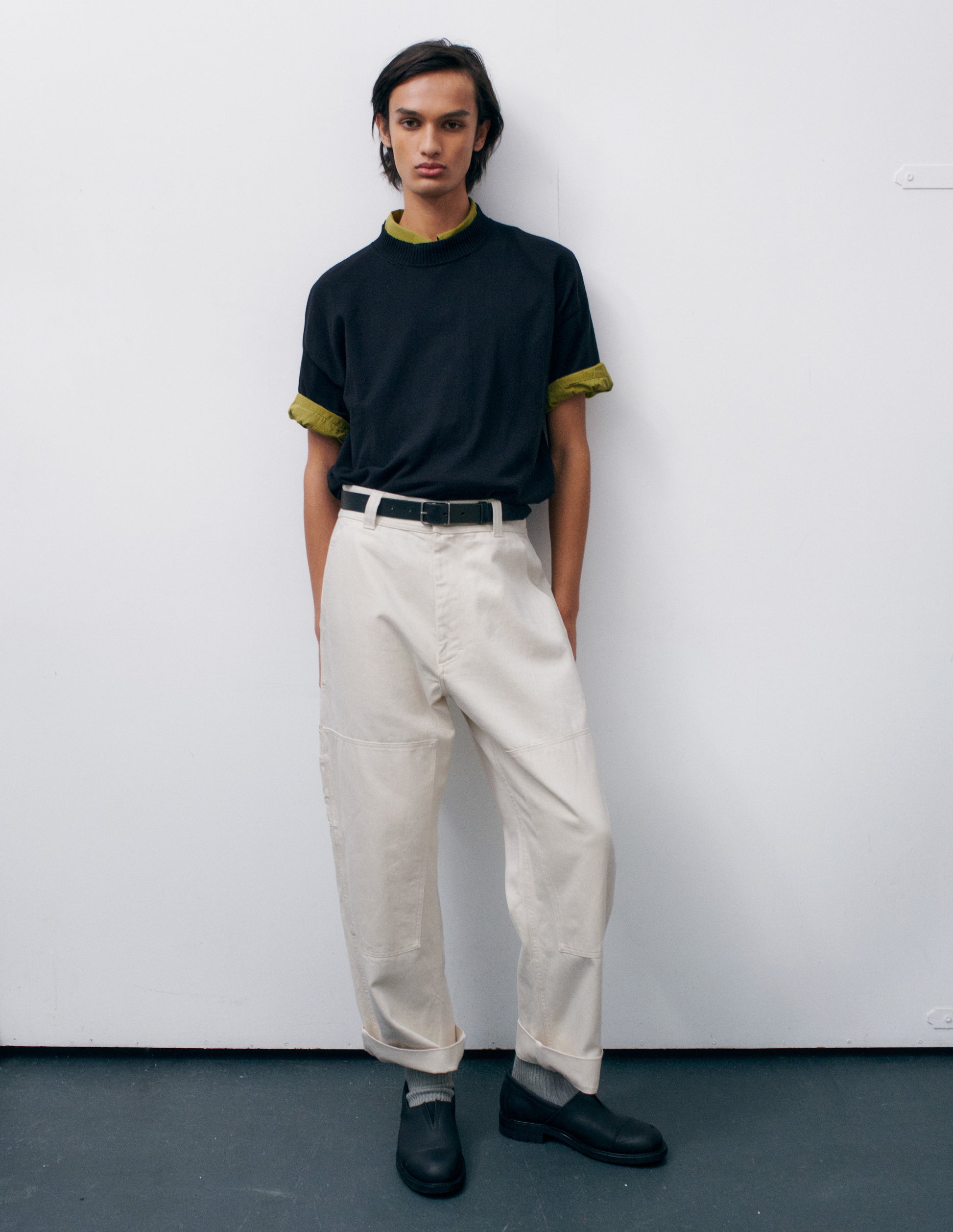 Full body image, front view of model wearing off white trousers with large knee patches, double tool pocket on the side leg and zip fly. Styled with green short sleeve shirt and black short sleeve jumper tucked into trousers, with black leather belt and black leather slip on shoes.