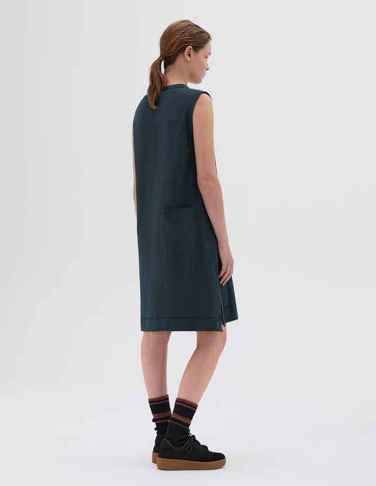 Full body image, back view of model wearing wearing blue-green regular cut knee length sleeveless and collarless dress with split side hem and patch back pocket, styled with black socks and black nubuck trainers.